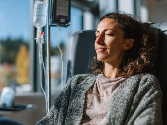 A serene woman relaxes in a comfortable chair while receiving an IV therapy treatment in a sunlit medical setting.